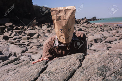 Man with a paperbag over his head is sitting on the beach